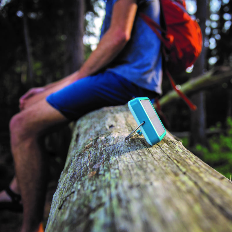 A BioLite SunLight 100 in teal colour, shown resting on it's kickstand upon a tree log next to someone sitting on the log in the woods.